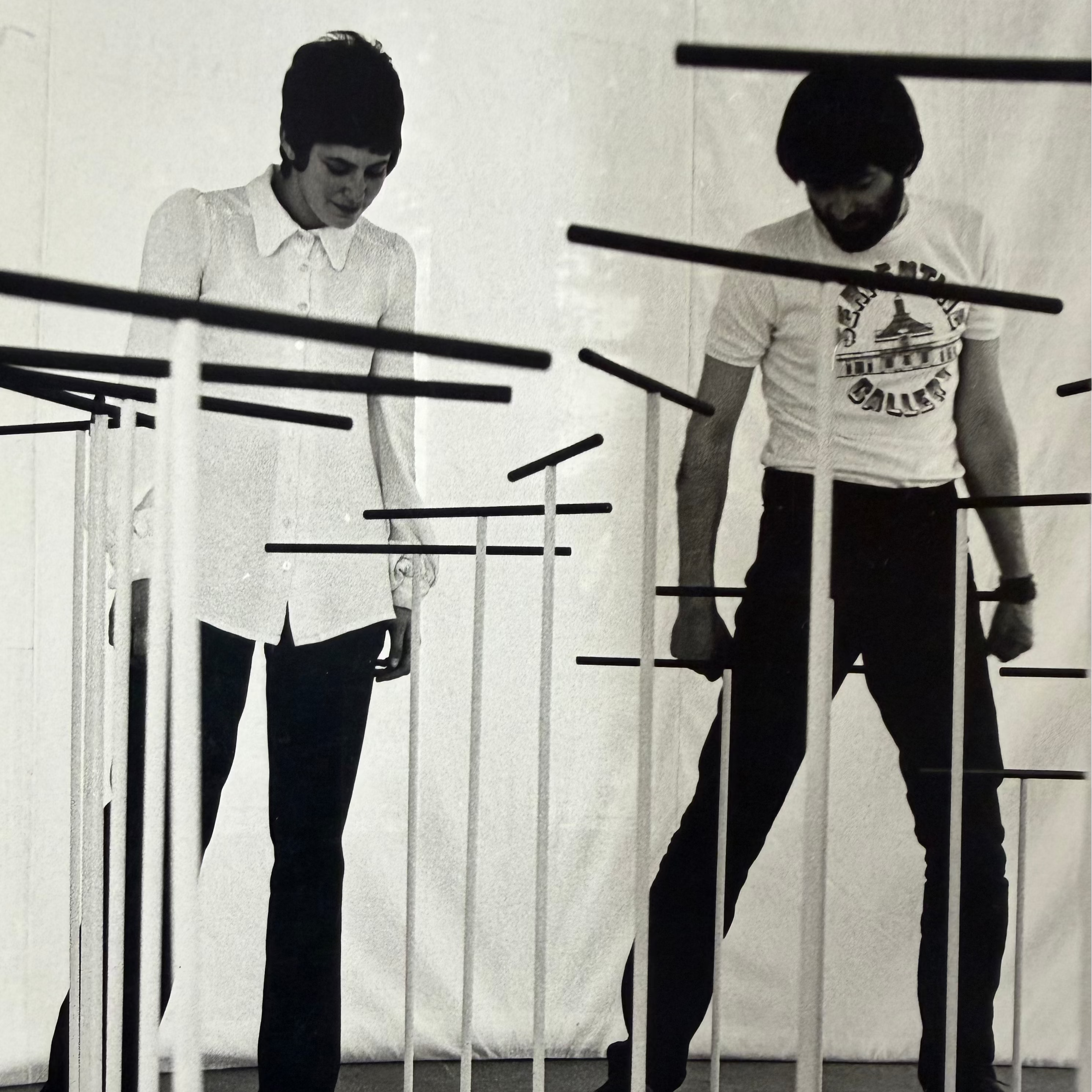 Black and white photograph of two people standing among tall black rods and circular platforms on the floor, posed in front of a draped white backdrop.